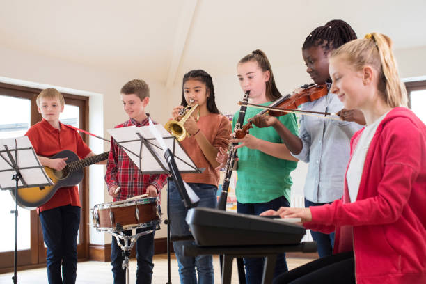 Children participating in a group music lesson, playing piano, guitar, trumpet, clarinet, violin, and drums while reading sheet music in a bright classroom.