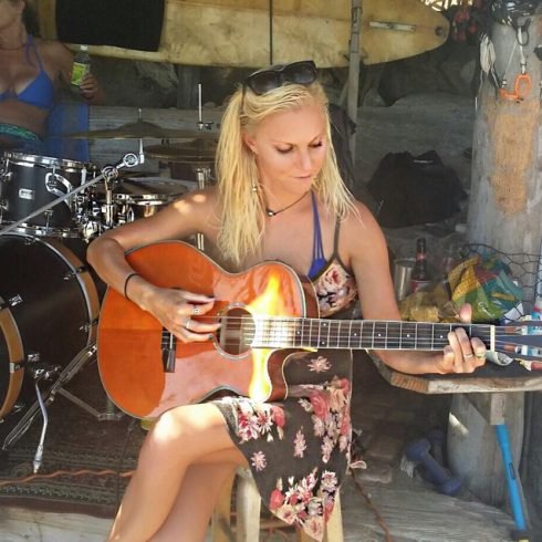Lindsay playing an acoustic guitar outdoors while seated near a drum set during a casual live music session.